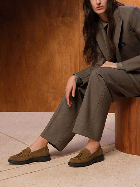 Two brown suede Spherica&trade; EC1 moccasins worn by a woman wearing a jacket and trousers on a wooden seat | GEOX | GEOX
